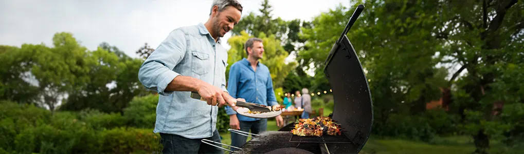 Un barbecue noir est au premier plan, avec des aliments en train de cuire. Des arbres et des personnes sont visibles en arrière-plan.