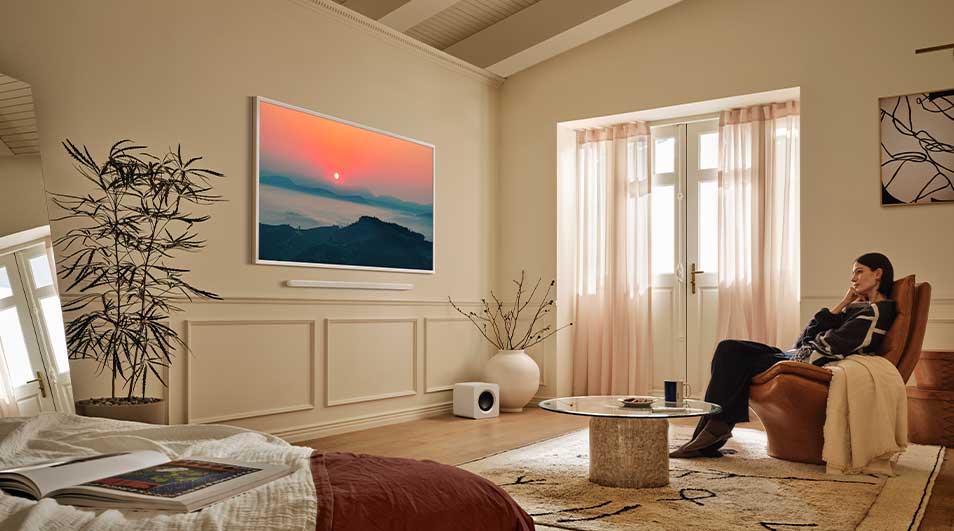 Een vrouw zit ontspannen op een fauteuil in een ruime, lichte woonkamer en kijkt naar een kunstwerk met een zonsondergang aan de muur.