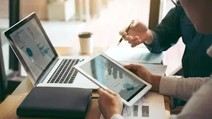 Une table montre un ordinateur portable, une tablette, des documents et une tasse à café. Les écrans affichent des graphiques.