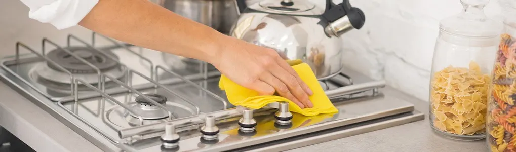 A stainless steel stovetop is being cleaned with a yellow cloth. A kettle and pasta jars are in the background.