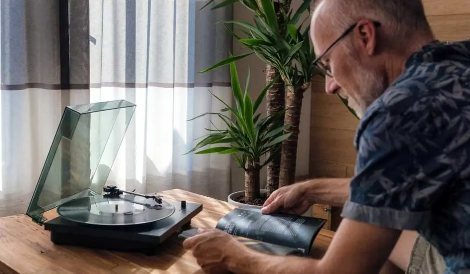 Een platenspeler met open deksel op een houten tafel, naast een plant. Een persoon leest een boek. Gordijnen op de achtergrond.