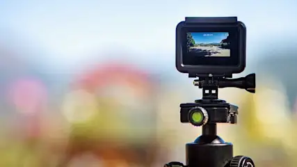 Een zwarte action camera op een statief, met een wazige achtergrond van lichtblauwe en roze tinten. Op het scherm is een landschap te zien.