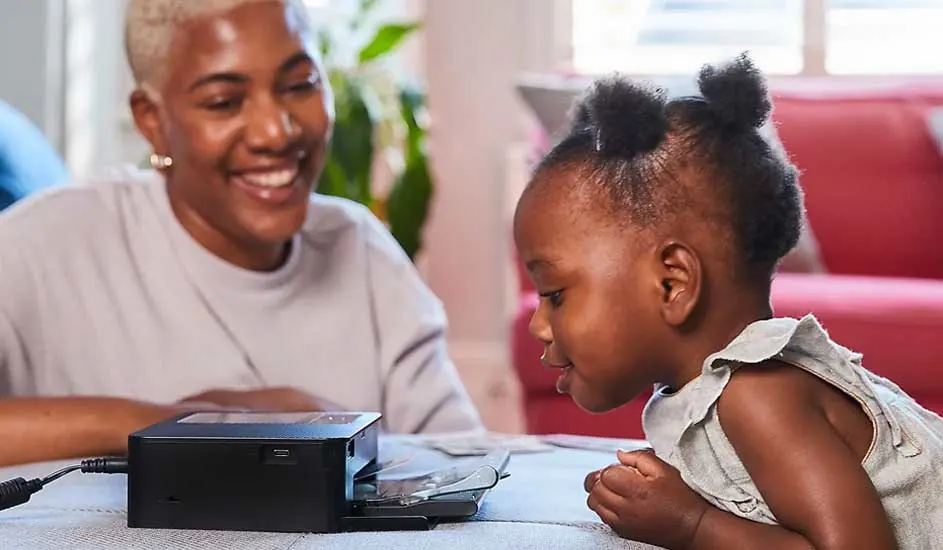 Une imprimante noire est posée sur une table grise. Une femme est assise en arrière-plan et une enfant regarde l'imprimante.