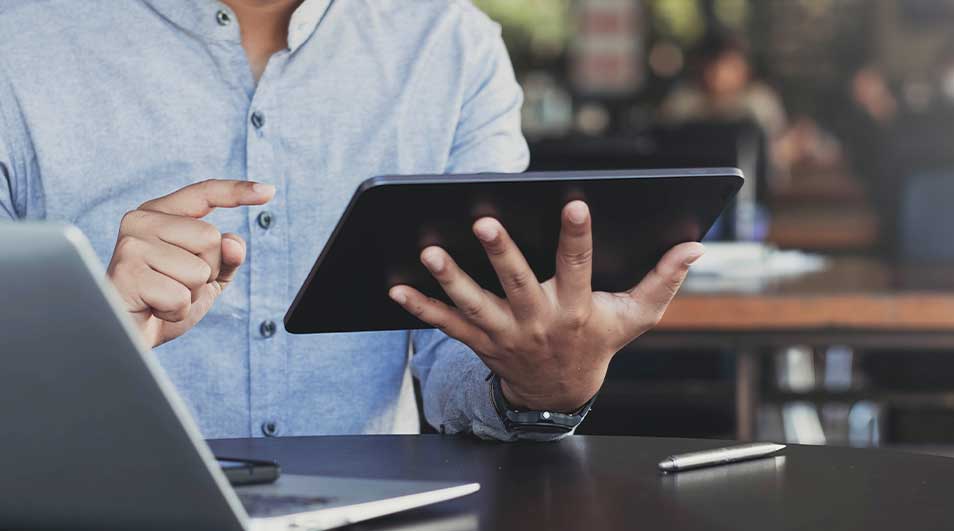 Een man gebruikt een tablet aan een bureau, met een laptop en pen erbij. Hij draagt een licht overhemd en werkt aandachtig.