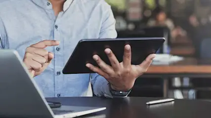 Een man gebruikt een tablet aan een bureau, met een laptop en pen erbij. Hij draagt een licht overhemd en werkt aandachtig.
