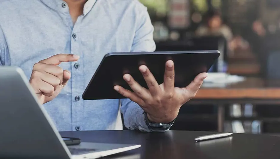 Un homme travaille sur une tablette à un bureau, avec un ordinateur portable et un stylo à côté. Il semble concentré et appliqué.