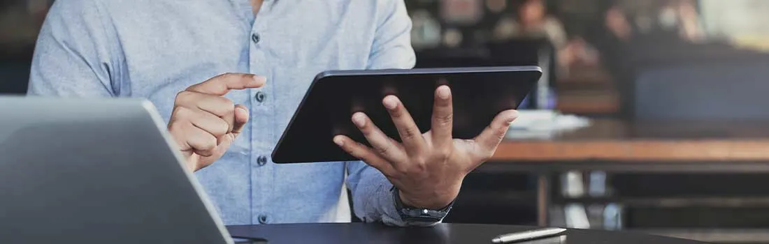 Een man gebruikt een tablet aan een bureau, met een laptop en pen erbij. Hij draagt een licht overhemd en werkt aandachtig.