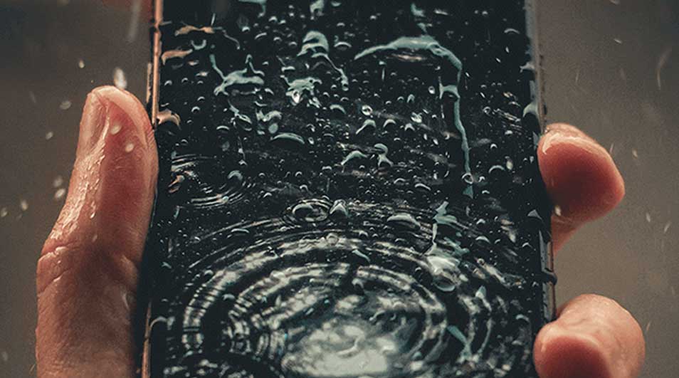 Un téléphone noir, couvert de gouttes d'eau, est tenu par deux doigts. L'eau forme des cercles concentriques sur l'écran.