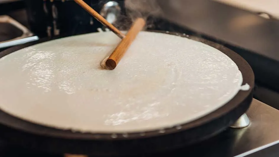 Une crêpe cuit sur une plaque noire, étalée avec un bâton en bois. Vapeur visible.