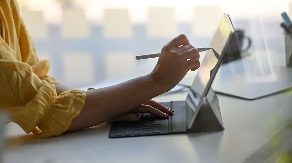 Een tablet op een bureau, een hand met een stylus op het scherm, een toetsenbord en een gele mouw.