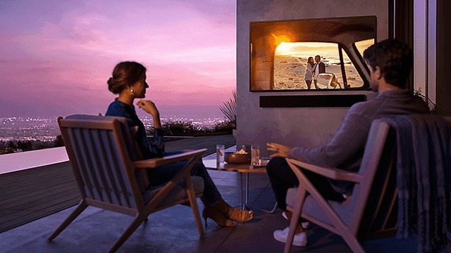 Un couple est assis dehors, regardant la télévision. Le ciel est rose et violet. Une table est entre les deux chaises.