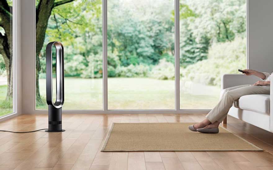 Dans un salon lumineux, une personne assise dirige une télécommande vers un ventilateur sans pales près de grandes fenêtres.