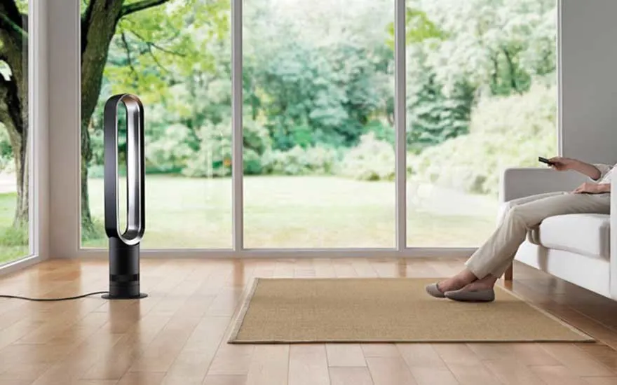 Dans un salon lumineux, une personne assise dirige une télécommande vers un ventilateur sans pales près de grandes fenêtres.
