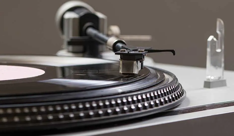 Zicht op een draaitafel met een zwarte vinylplaat en een toonarm met naald in focus. Achtergrond toont een wazig object.