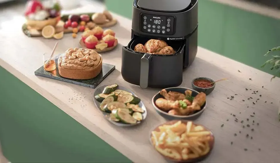 Dit is een Philips airfryer op een keukenblad met kip, friet, courgette, taart en muffins eromheen, alles kleurrijk gepresenteerd.
