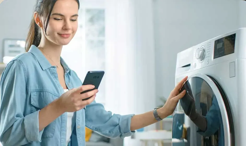Een vrouw staat glimlachend bij een wasmachine, houdt haar smartphone vast en raakt de deur aan in een lichte, moderne wasruimte.