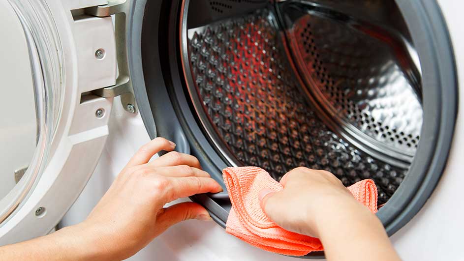 Une personne nettoie le joint gris d'un lave-linge blanc avec un chiffon orange. Le tambour est en métal argenté perforé.