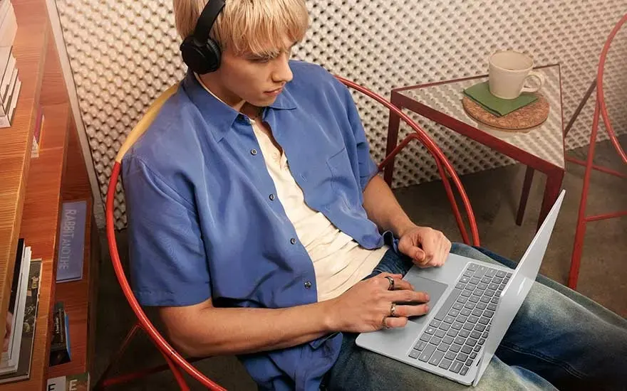 Een persoon met kort blond haar werkt op een laptop in een moderne werkruimte, draagt een blauwe overhemd en koptelefoon.