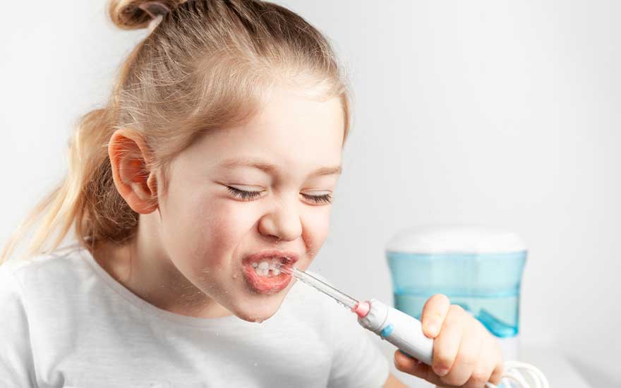 A young girl uses a water flosser, eyes closed and face splashed with water while cleaning her teeth.