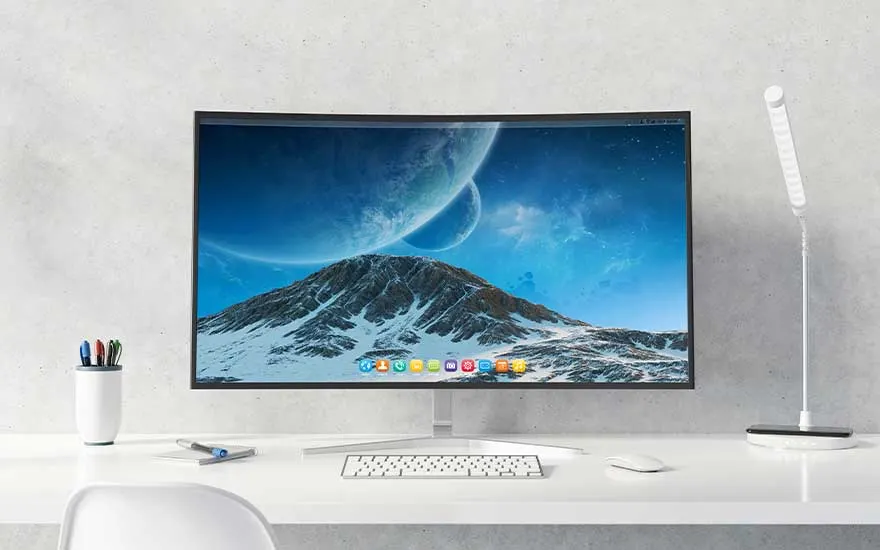 Een gebogen monitor toont een besneeuwde berg onder een blauwe hemel met planeten. Een wit bureau heeft een toetsenbord, lamp en pennen.