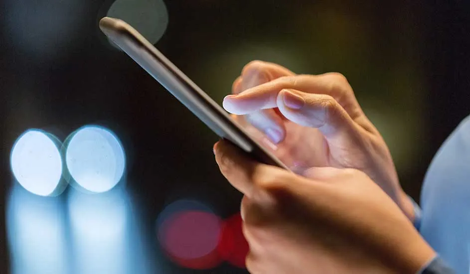 Main tenant un téléphone, doigt touchant l'écran. Arrière-plan sombre avec lumières floues.