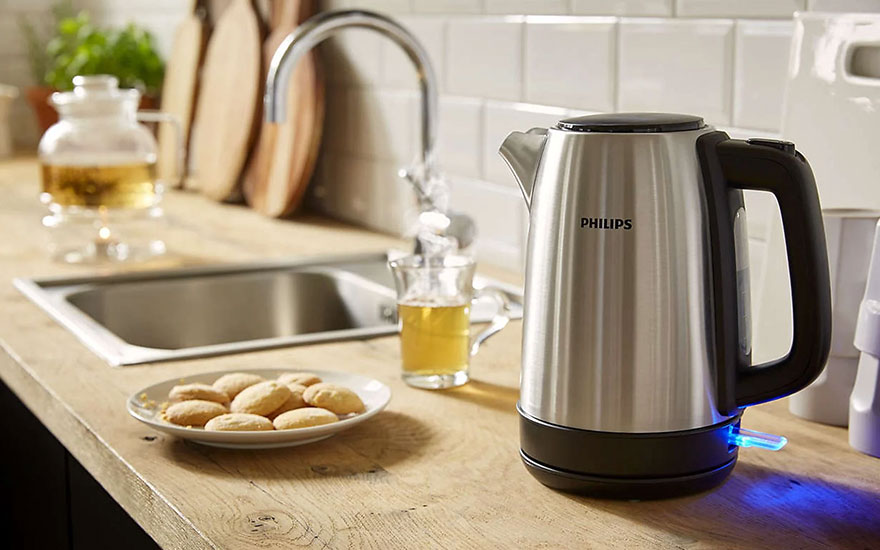Een zilverkleurige Philips waterkoker staat op een houten aanrecht, naast een gootsteen en koekjes op een bord.