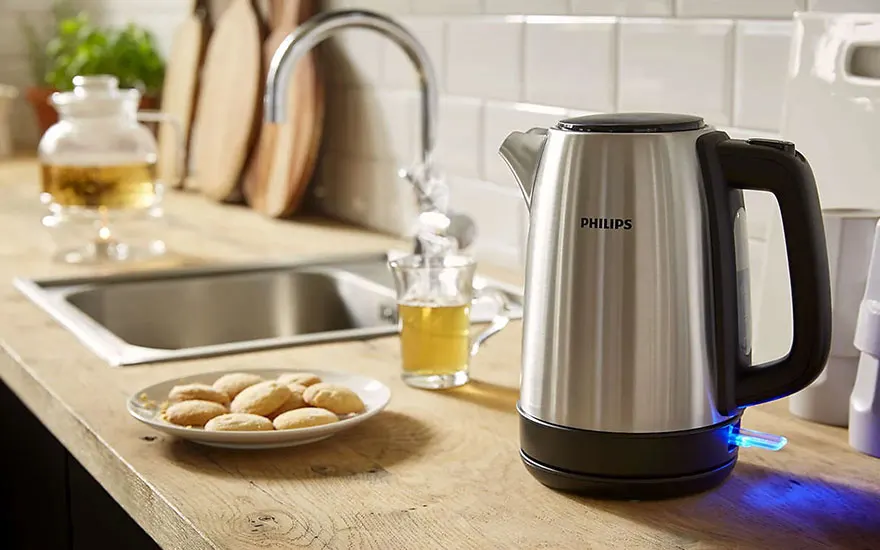 Een zilverkleurige Philips waterkoker staat op een houten aanrecht, naast een gootsteen en koekjes op een bord.