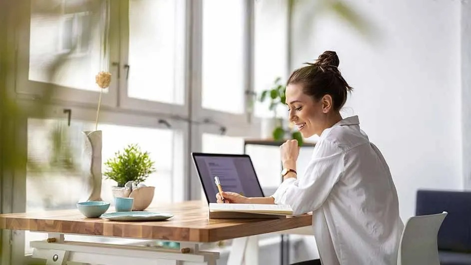 At a desk with plants, a laptop, and dishes, a pen hovers over an open notebook. A bright window is in the background.