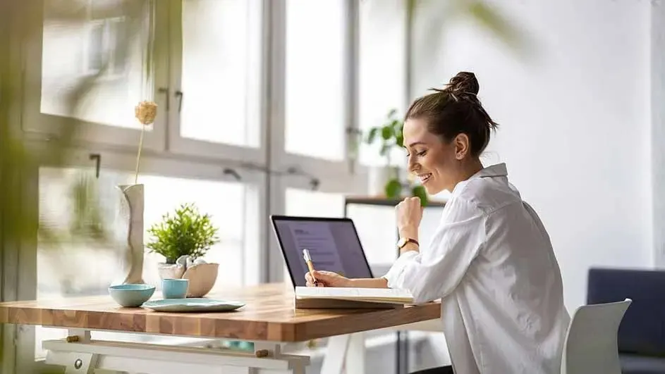 At a desk with plants, a laptop, and dishes, a pen hovers over an open notebook. A bright window is in the background.