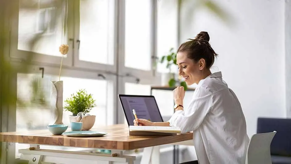 Een houten bureau met een laptop, notitieboek en kantoorartikelen staat voor een groot raam in een lichte ruimte.