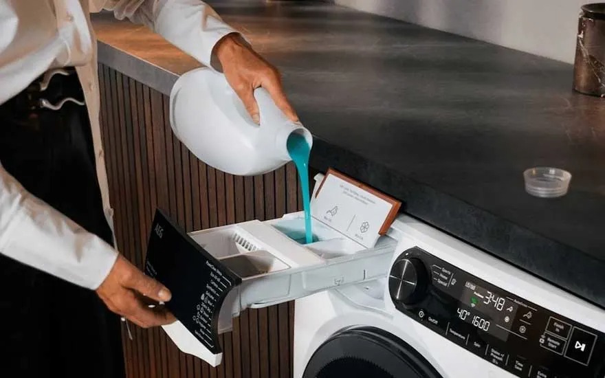 Iemand giet blauw vloeibaar wasmiddel in het doseerbakje van een witte AEG wasmachine in een moderne wasruimte.