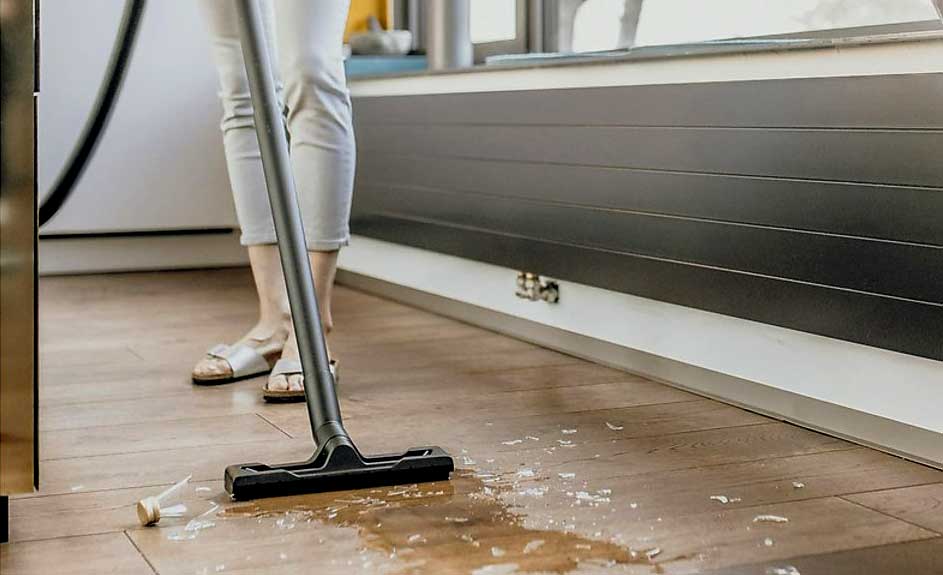 Aspirateur noir sur parquet avec liquide clair et bouchon. Fenêtre et plinthe grise en arrière-plan.
