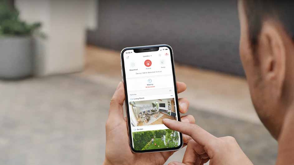 Een persoon houdt een smartphone vast met een app voor huisbeveiliging, met beelden van een woonkamer en tuin.