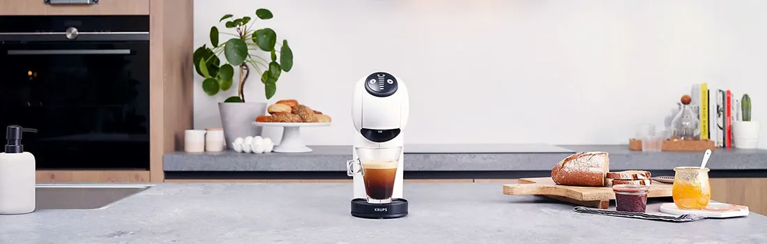 Plan de travail de cuisine avec machine à café blanche, tasse de café, pain et confiture. Un four et des plantes sont visibles.