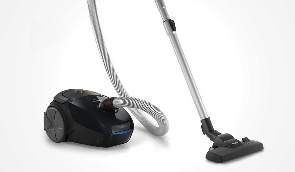 Black cylinder vacuum with grey hose and metal tube attached to wide floor head, positioned at an angle on a smooth light background.