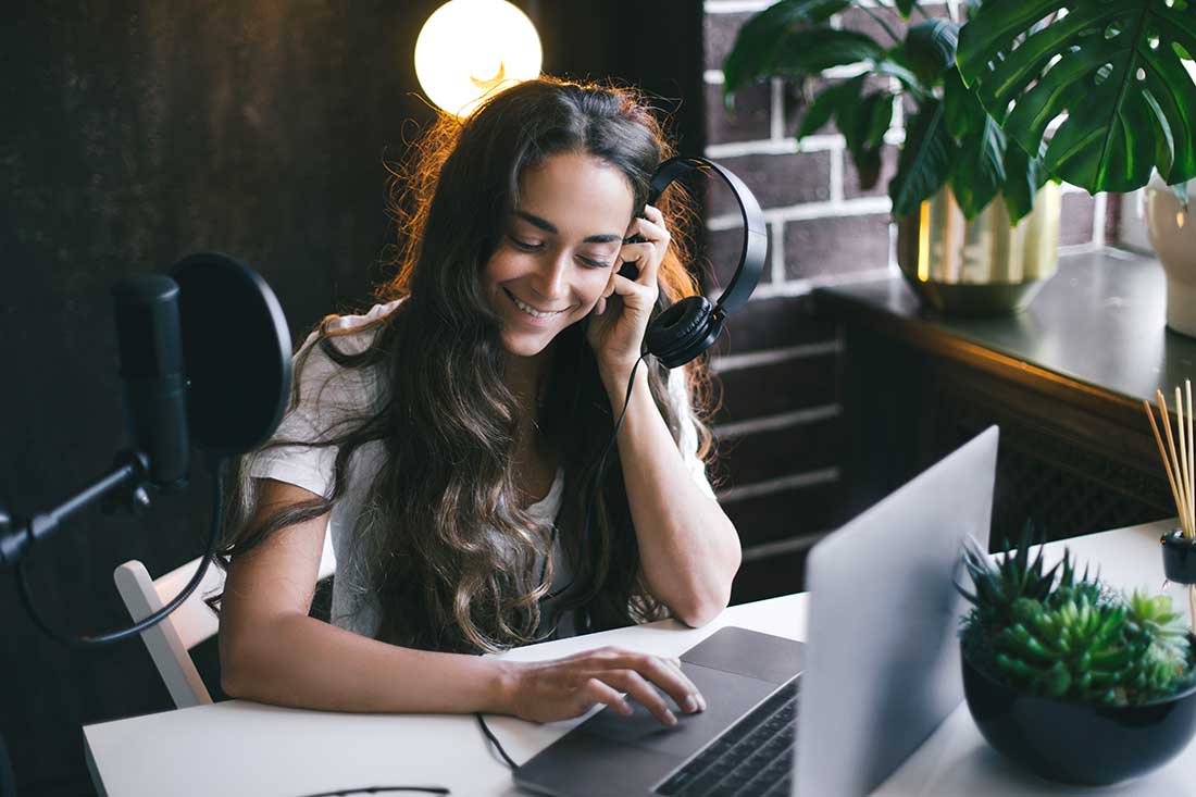Une personne utilise un ordinateur portable sur un bureau blanc avec un microphone et des écouteurs à proximité. Une plante est visible.