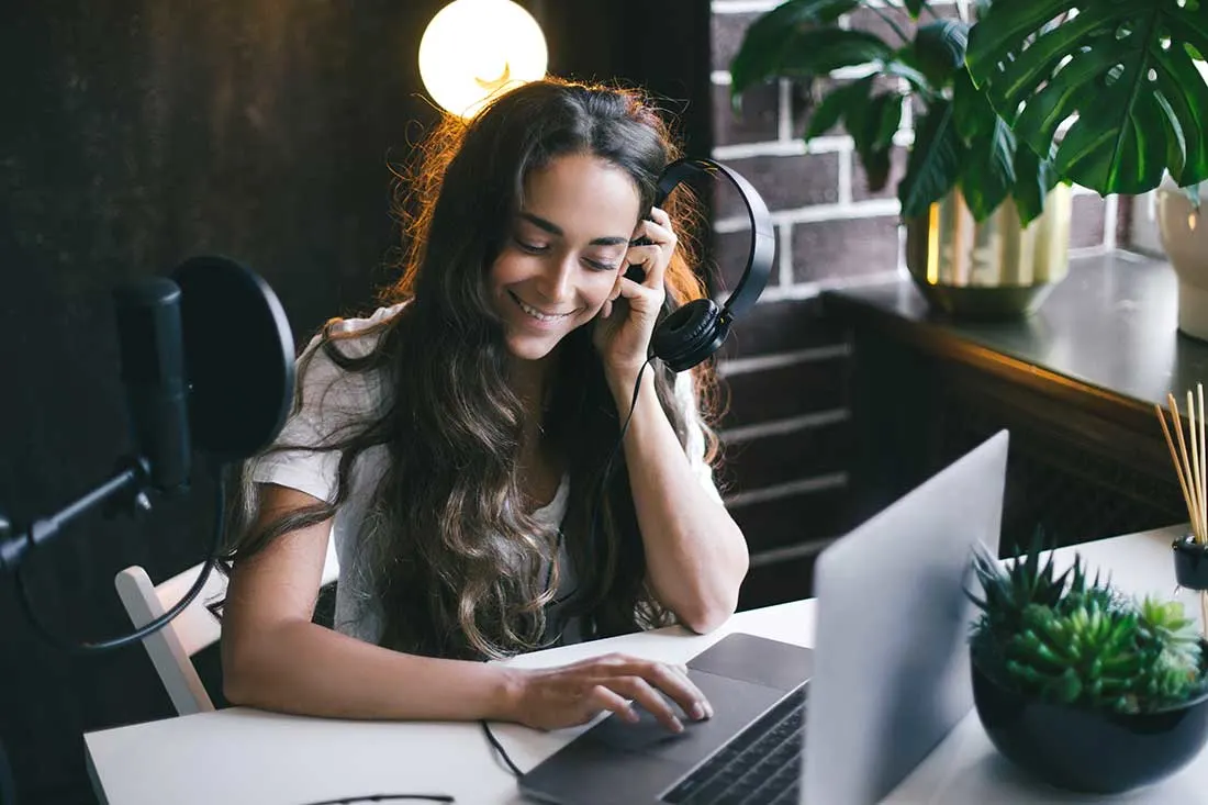 Une personne utilise un ordinateur portable sur un bureau blanc avec un microphone et des écouteurs à proximité. Une plante est visible.