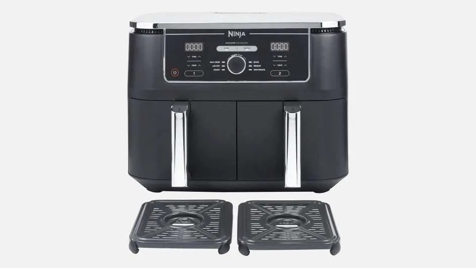 Een zwarte Ninja airfryer met twee lades en zilveren handvatten staat centraal, met twee losse grillplaten ervoor op een lichte achtergrond.