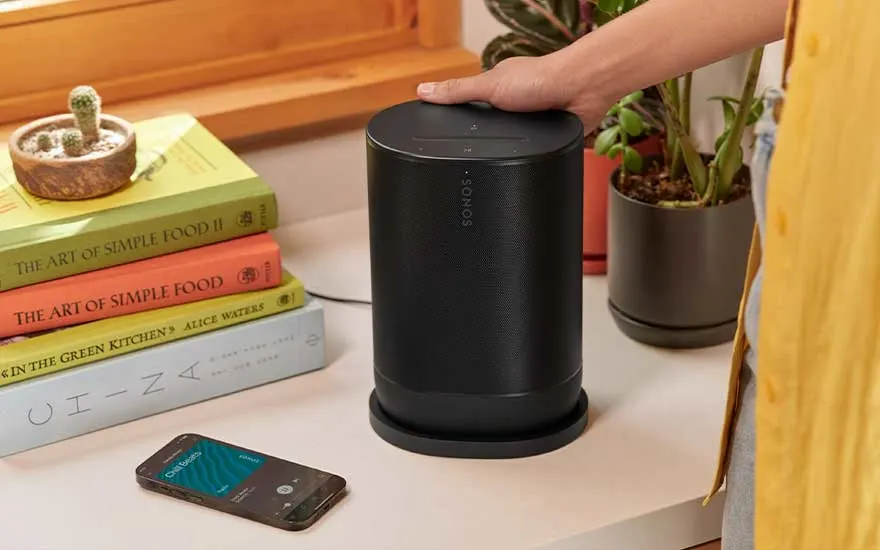 Zwarte Sonos-luidspreker op tafel naast boekenstapel, smartphone en planten. Hand raakt de bovenkant van de luidspreker aan.