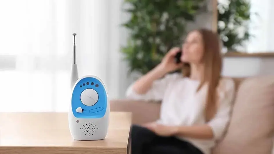 Un babyphone blanc et bleu est posé sur une table en bois. Un autre appareil avec une antenne est visible en arrière-plan.