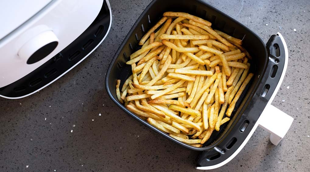 Vue de dessus d'une friteuse à air noir remplie de frites dorées sur un comptoir gris près d'un appareil blanc.