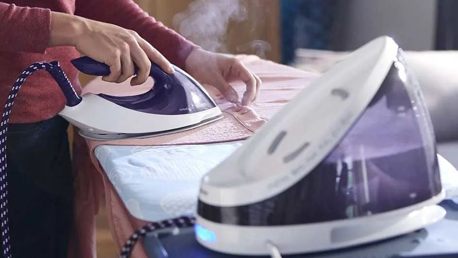 Een stoomstrijkijzer, wit en paars, strijkt kleding op een blauwe strijkplank met stoom. Een stoomgenerator staat ernaast.