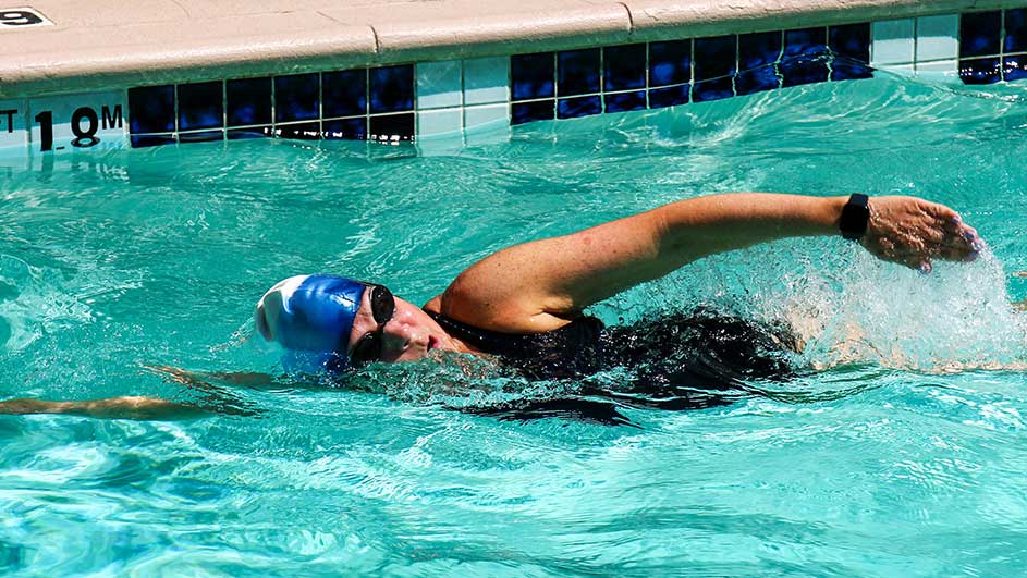 Une personne nage dans une piscine turquoise. Elle porte un bonnet de bain bleu et des lunettes de natation noires. On voit le bord de la piscine.