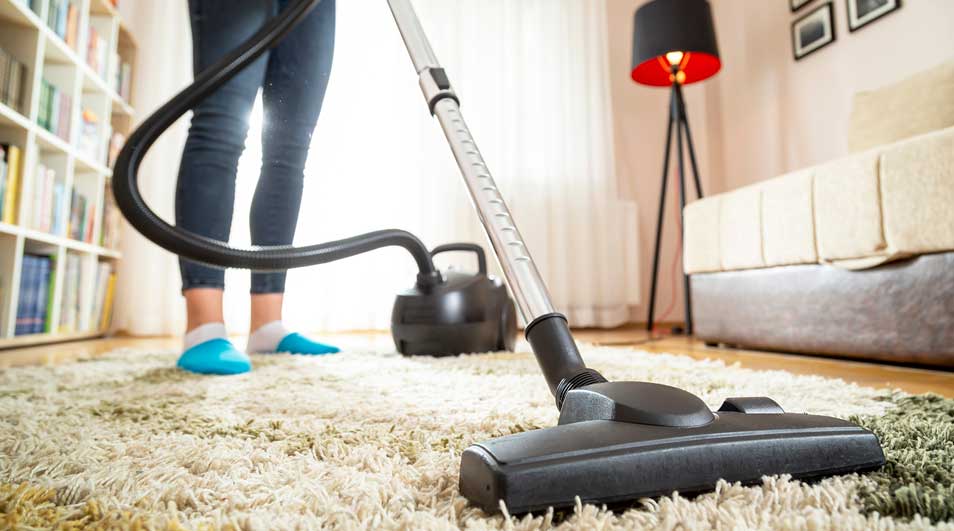 Aspirateur noir sur tapis beige, tube noir visible. Bibliothèque, lampe et canapé en arrière-plan.