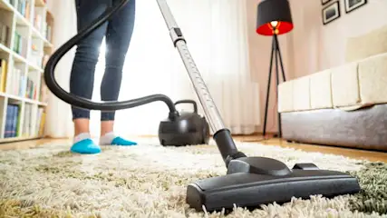 Aspirateur noir sur tapis beige, tube noir visible. Bibliothèque, lampe et canapé en arrière-plan.