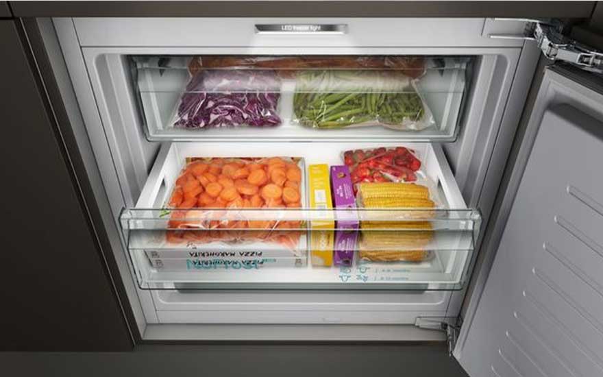 Een geopende witte vriezer gevuld met groenten, fruit en pizza's in plastic verpakkingen in transparante lades.