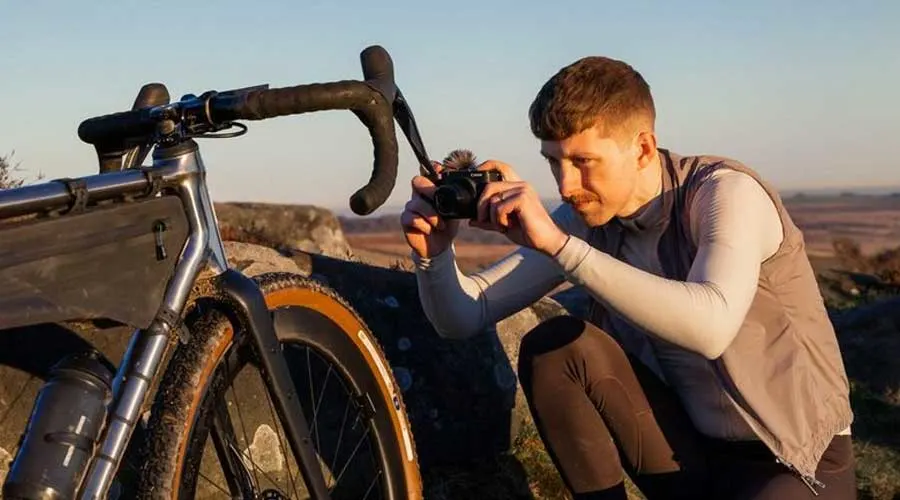 Cyclist crouching by a gravel bike, taking a close-up photo with a compact camera at golden hour outdoors.
