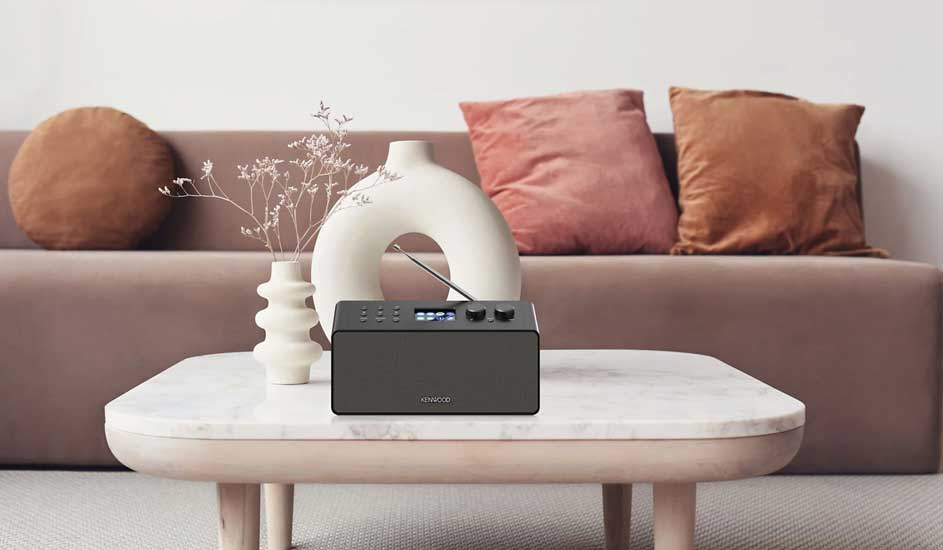 Donkergrijze Kenwood radio op een marmeren tafel. Achtergrond: bruine zetel met kussens en decoratieve vazen.
