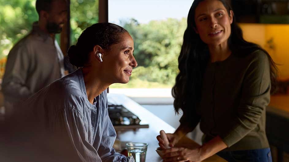 Indoor shot. Woman with earbud in blue shirt, glass of water on counter. Another woman in green sweater. Window with trees behind.