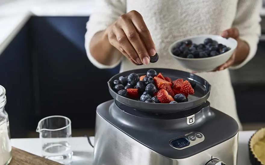 Een persoon bereidt een gezond dessert en strooit blauwe bessen over aardbeien in een schaal op een moderne keukenmachine.